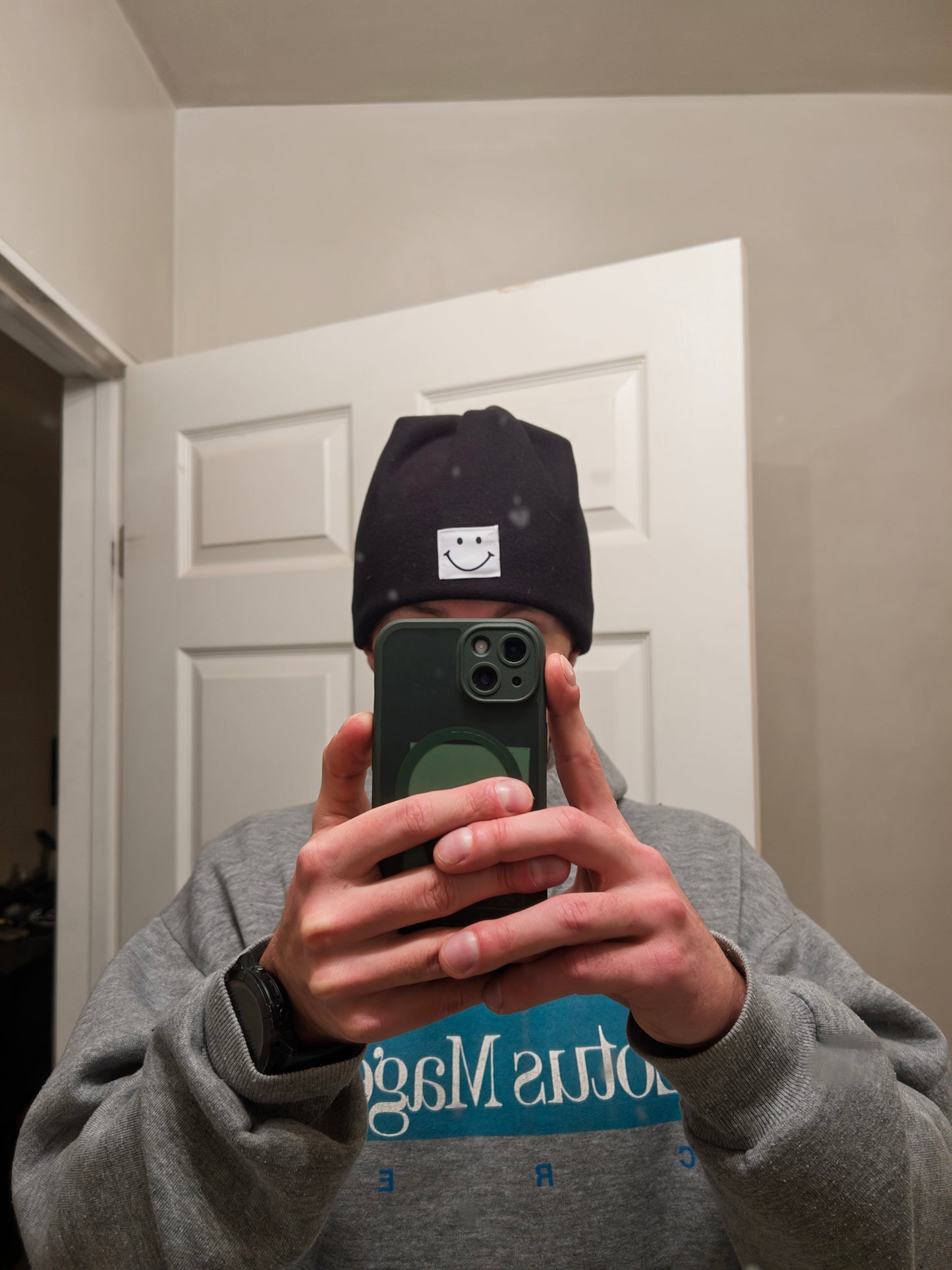 Person wearing a smiley face cap and gray sweatshirt taking a mirror selfie.