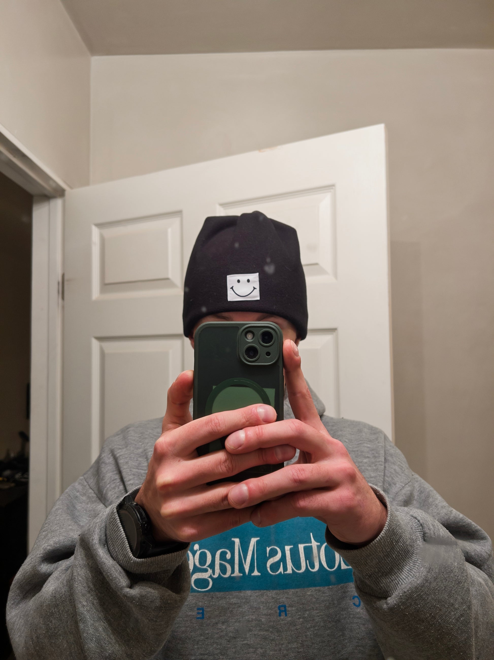 Person wearing a smiley face cap and gray sweatshirt taking a mirror selfie.